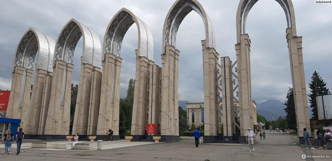 Архитектурная композиция из арок в городе, сквер с зелеными насаждениями и горы на фоне, люди гуляют по площади