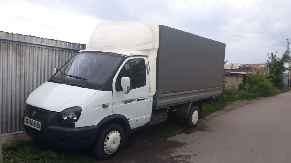 Ақ түсті жүк фургоны, сұр шатырмен, тар жолда металл қоршаудың жанында паркталған.