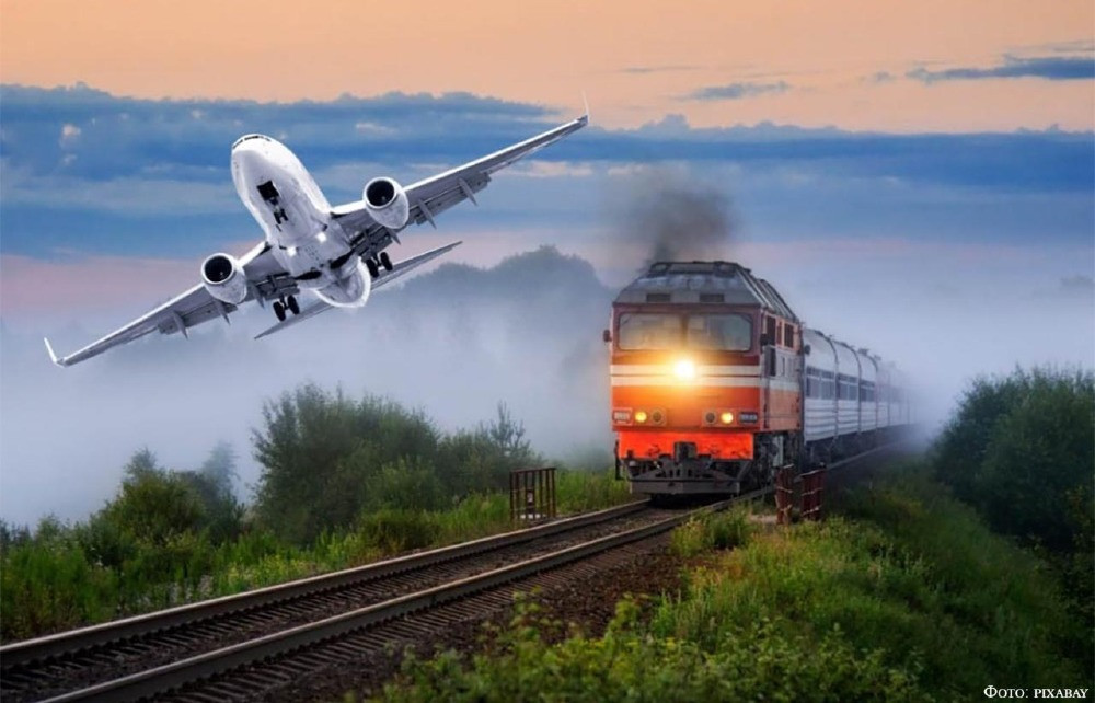 Поезд движется по рельсам в туманном пейзаже, в небе над ним летит самолет