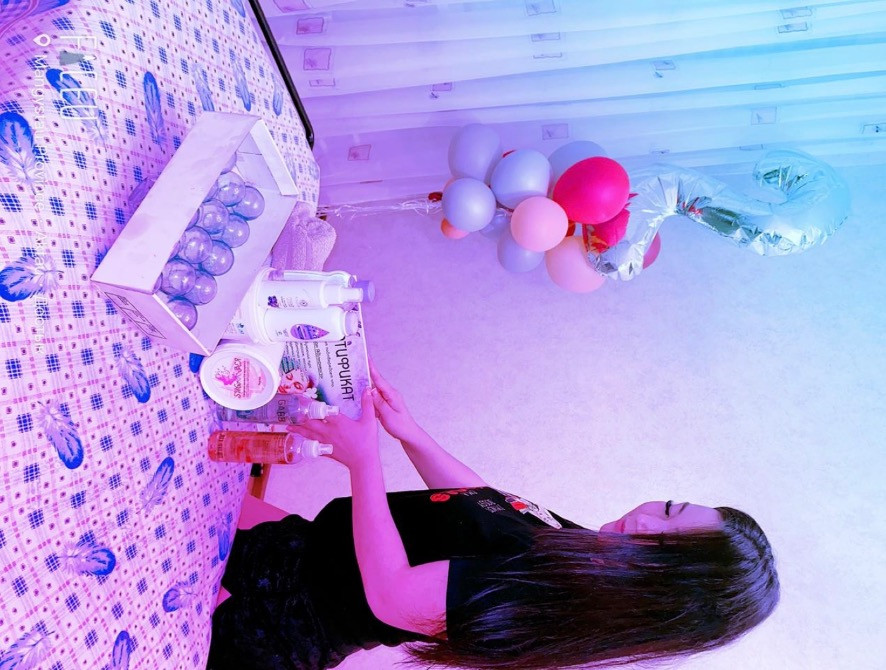 Қыз үстелдің жанында тұр, үстелде косметикалық құралдар мен шариктері бар қаптама орналасқан. Артқы планда түрлі-түсті шарлар көрініп тұр