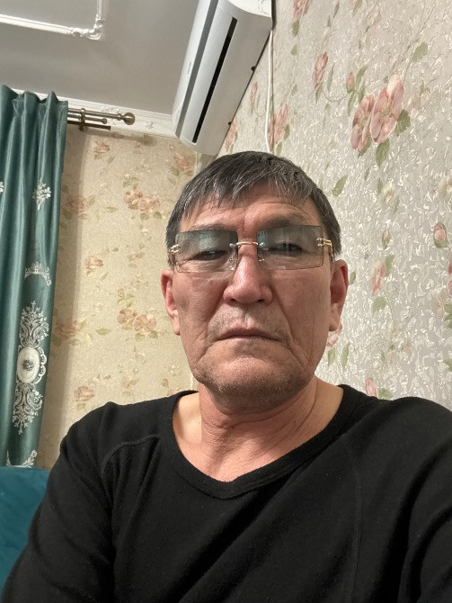 Қара киімдегі ер адамның портреті түрлі-түсті қағаздар мен перделердің фонында. Ер адам қысқа шашты және көзілдірікті.