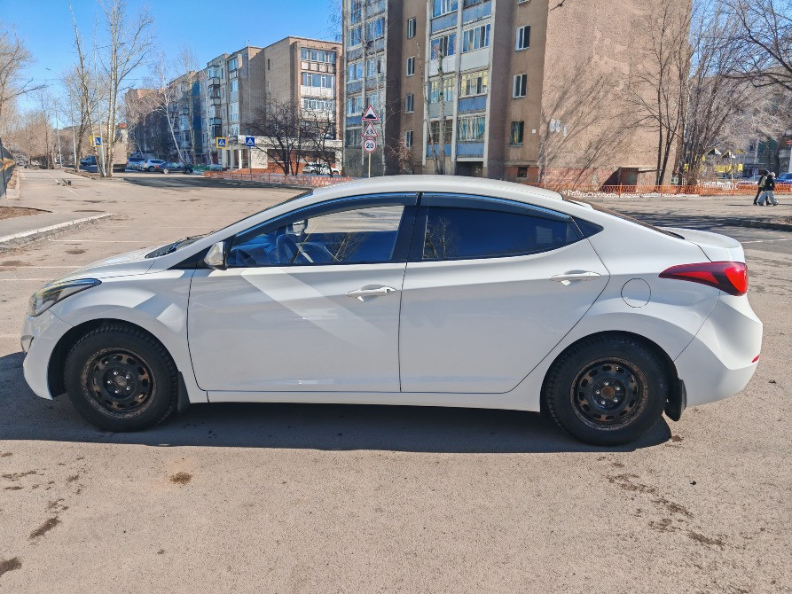 Ақ Hyundai Elantra автокөлігі көп қабатты үйлер фонында бокстан көрініс