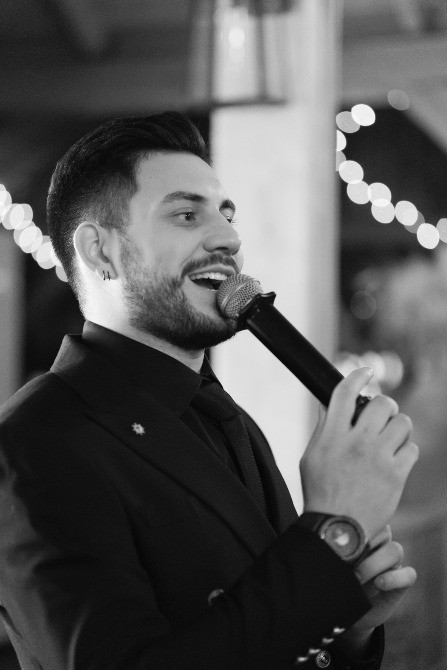 Мужчина с микрофоном выступает на сцене, одет в черный костюм