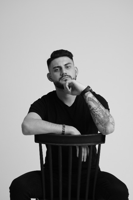 Мужчина с короткой стрижкой и татуировками сидит на стуле, смотрит вдаль