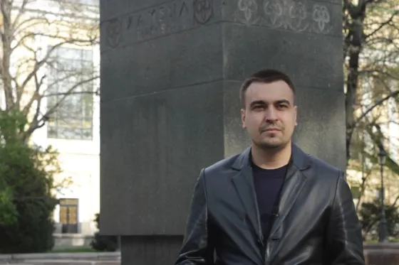 Мужчина в кожаной куртке стоит на фоне памятника, окружающего зелени
