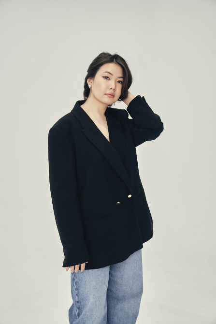 Үлкен пиджак пен джинса киген әйел, ашық фонда поза беріп тұрған