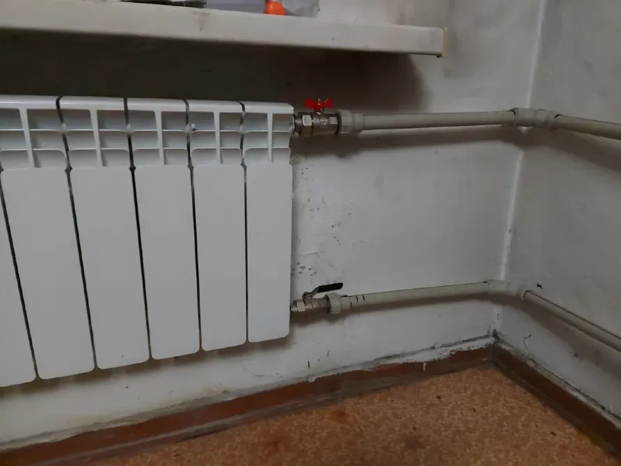 Бөлменің бұрышына орнатылған құбырлары бар жылыту радиаторы, көрінетін қосылыстар және вентиль