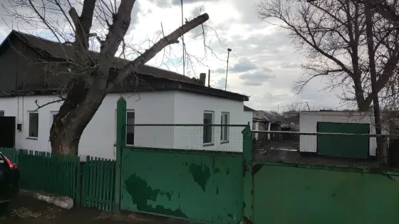 Дом с белыми стенами и черной крышей, огражденный зеленым забором, рядом с деревом и другим зданием на заднем плане