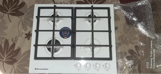 Газовая плита с четырьмя конфорками и черной решеткой на белой поверхности. Кнопки управления расположены внизу. Фон с цветочным узором
