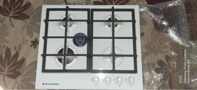Газовая плита с четырьмя конфорками и черной решеткой на белой поверхности. Кнопки управления расположены внизу. Фон с цветочным узором
