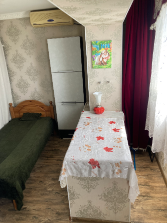 Уютная комната с односпальной кроватью, холодильником и столом, оформленная в светлых тонах