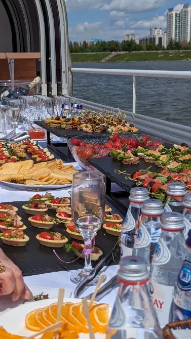 Стол с закусками и напитками на фоне реки, украшенный фруктами и бокалами