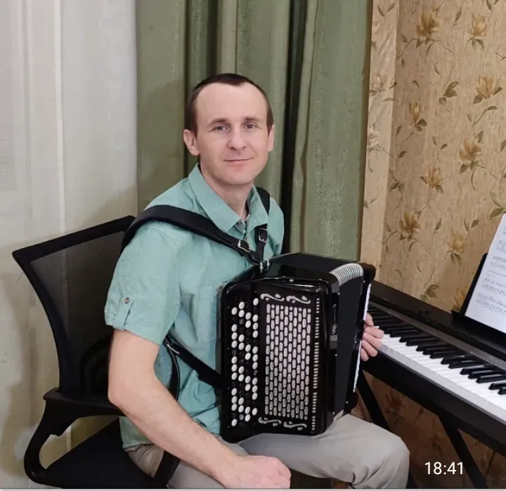 Игрок на баяне позирует в одежде, сидя рядом с фортепиано