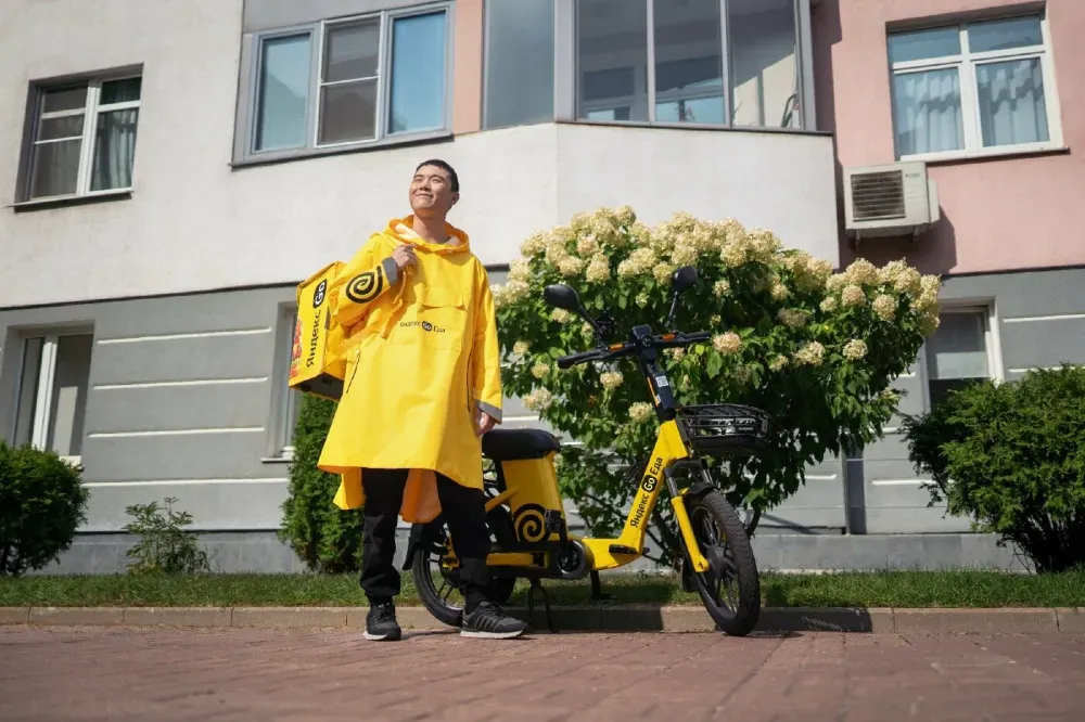 Человек в желтом плаще рядом с электрическим велосипедом Яндекс