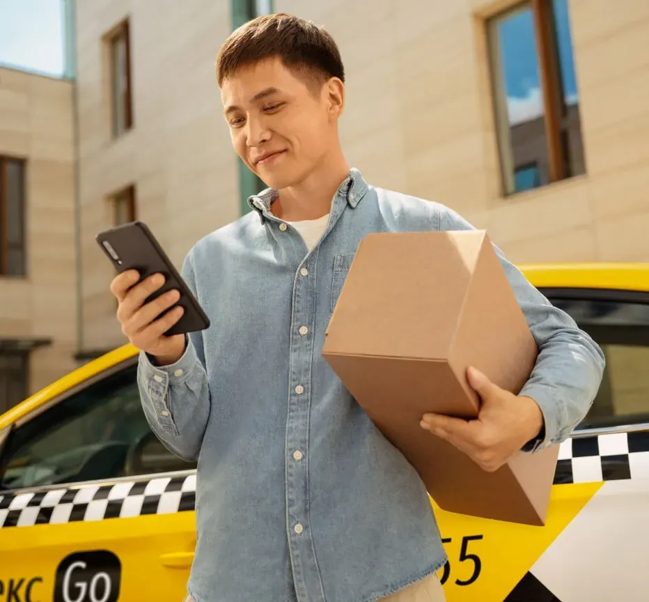 Мужчина с коробкой и телефоном на фоне желтой машины Яндекс
