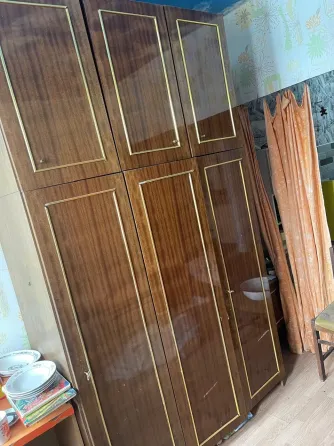 Деревянный шкаф с золотистой отделкой, расположенный в уголке комнаты, на фоне текстильных занавесей и различных предметов интерьера