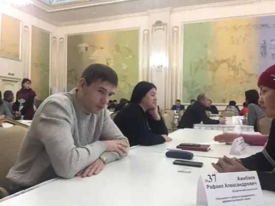 Сұр свитер киген адам талқылау кезінде үстелге мұқият қарап отыр, бөлмеде көп адам бар