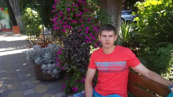 Қызыл футболка киген жас жігіт гүлдеген өсімдіктердің арасында орындықта отыр