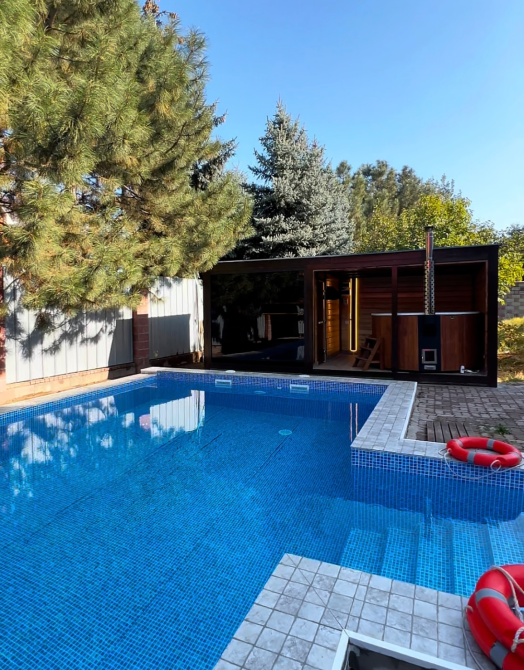 Бассейн с уютной зоной для отдыха и деревянной баней в окружении зелени