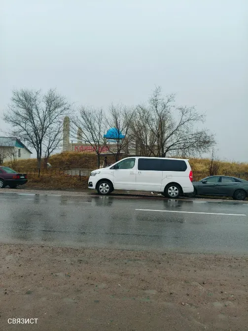 Ақ минивэн дымқыл жолда, артында ағаштар