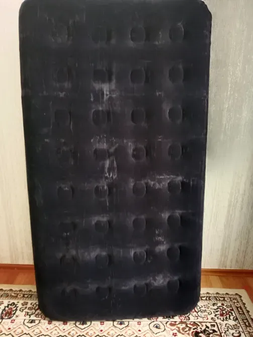 Надувная кровать, свернутая и стоящая вертикально на фоне ковра