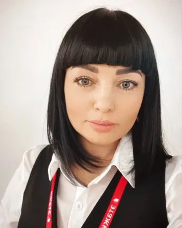 Портрет молодой женщины с короткой черной стрижкой и выразительными глазами. Она одета в белую рубашку и черный жилет, на шее у нее красный значок. В 