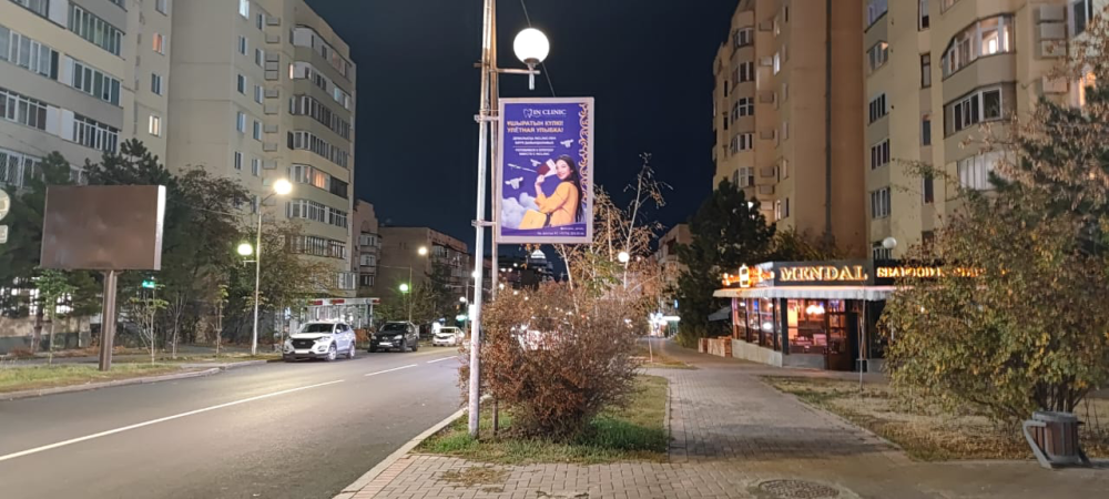 Тұрғын ауданда түнгі уақытта медициналық қызметтер туралы жарнамалық щитпен көше