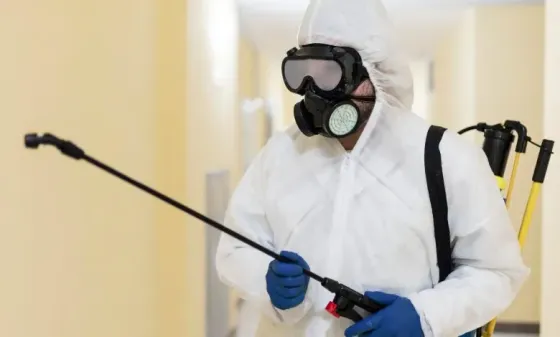 Работник в защитной одежде и маске, использующий распылитель для дезинфекции помещений
