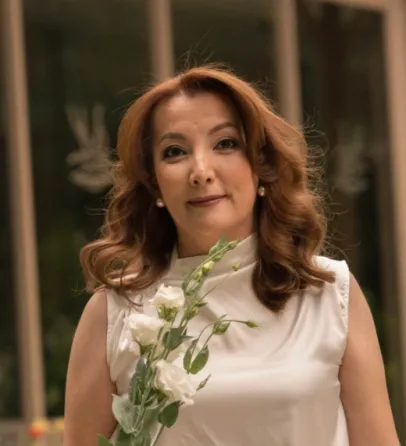 Женщина с длинными волнистыми волосами, одетая в светлую блузу, стоит на фоне стеклянной стены, держа в руках белые цветы