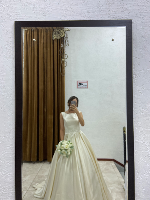Невеста в свадебном платье, стоящая перед зеркалом, с букетом в руках и отражением в зеркале