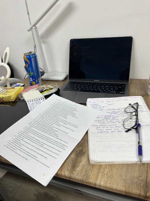 Ноутбук, блокнот, құжаттар, көзілдірік және канцелярлық құралдармен жабдықталған жұмыс үстелі.