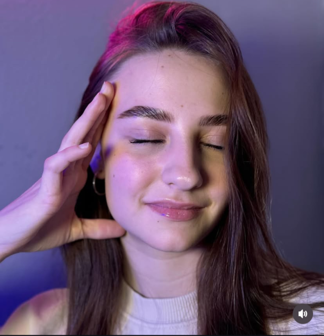 Девушка с закрытыми глазами, прикасается к своему лицу и выглядит расслабленной