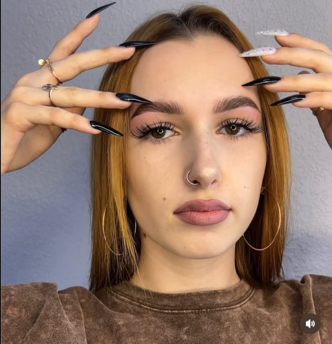 Молодая женщина с длинными ногтями и выразительным макияжем смотрит прямо в камеру, при этом прикоснувшись к лицу