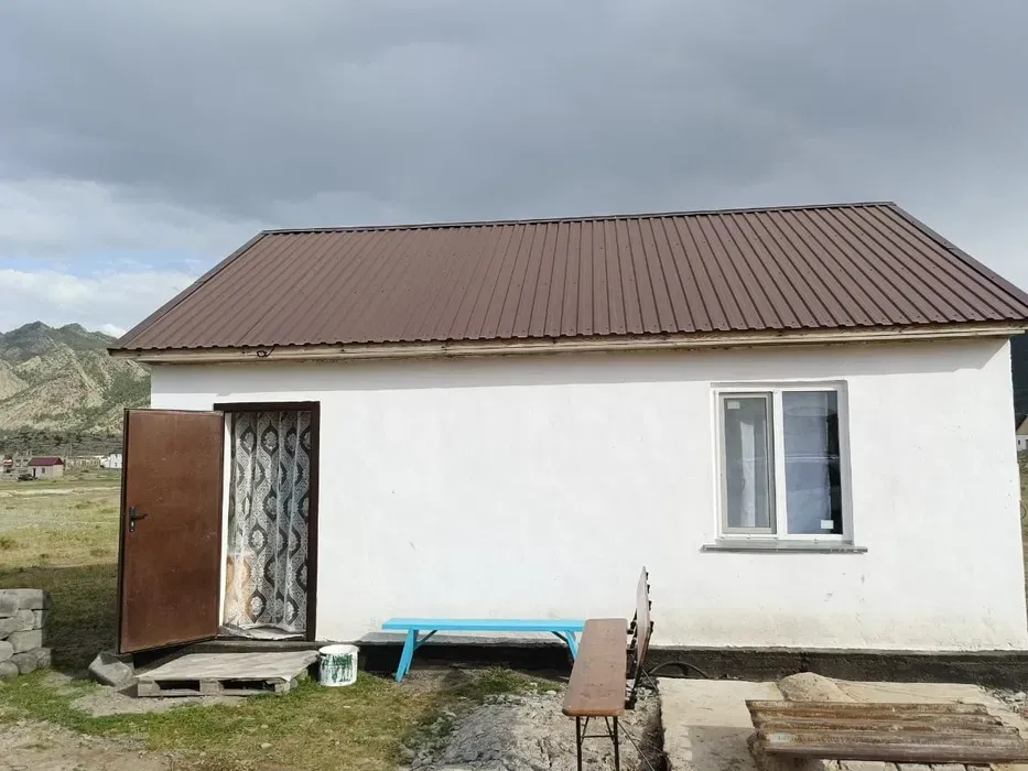 Металл шатырлы және ағаш есікті үй, бұлтты аспанның фонында жасыл төбелердің арасында орналасқан.