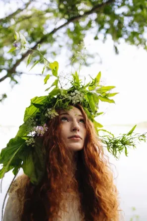 Ұзын қоңыр шашты, басында жасыл гүл шоқымен табиғат фонында жоғары қарай қараған қыз.