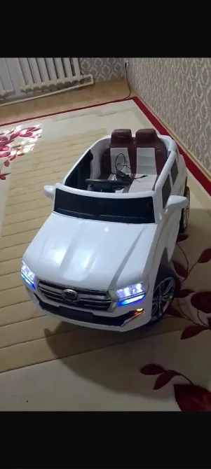 Ақ түсті ашық төбелі, қызыл орындықтары мен жарқын фарлары бар кішкентай ойыншық машина, кілемнің үстінде тұр.