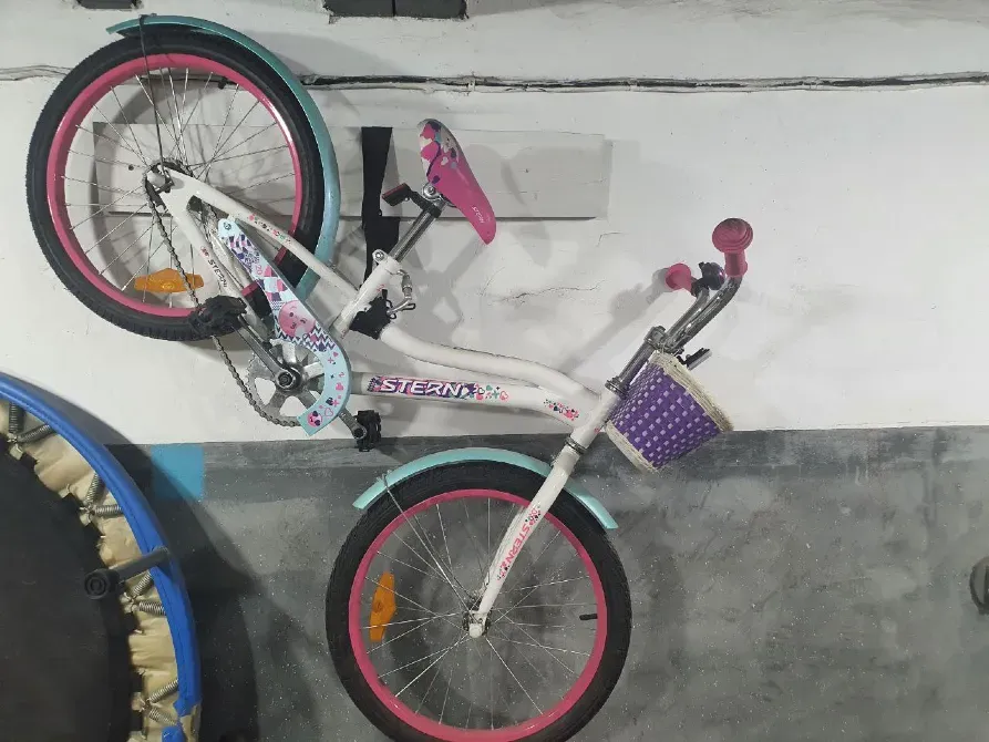 Детский велосипед с розовыми деталями и корзиной, висящий на стене гаража