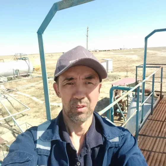 Мужчина на фоне промышленного оборудования в пустынной местности, одет в рабочую одежду и кепку