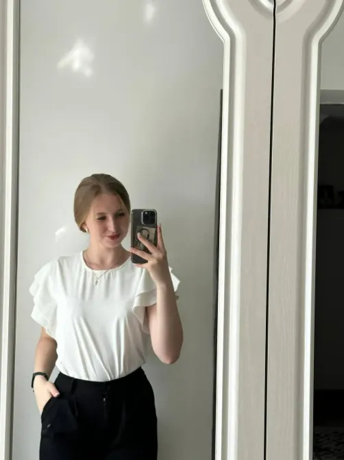 Девушка в белой блузке с рюшами позирует перед зеркалом, держит телефон и улыбается