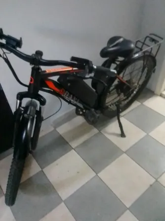 Үлкен дөңгелекті электрлік велосипед, рамада орнатылған аккумулятормен. Терезелері мен үстелі бар бөлмеде орналасқан.