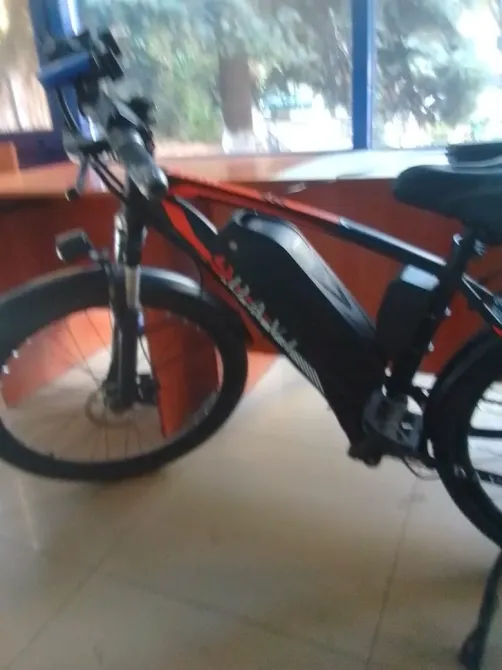 Үлкен дөңгелекті электрлік велосипед, рамада орнатылған аккумулятормен. Терезелері мен үстелі бар бөлмеде орналасқан.