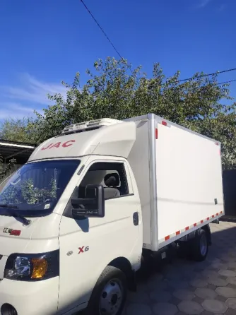 Грузовик JAC X6 с крытым кузовом на фоне голубого неба и зелёных деревьев