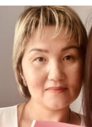 Қысқа шашты, ашық түсті шашы бар, жеңіл макияжбен боялған әйел камераға қарай қарап тұр. Ол күлімсіреп, қолында картон ұстаған.