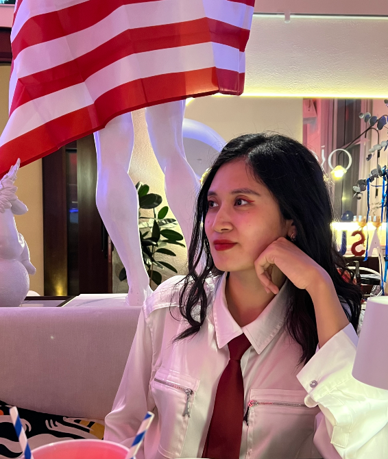 Девушка с длинными черными волосами в белой рубашке и красном галстуке, сидит в кафе с декоративным элементом на фоне