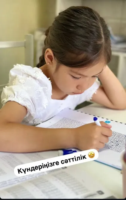 Қыз бала мәтін жазу үстінде, үстелдегі материалдарды мұқият қарап отыр