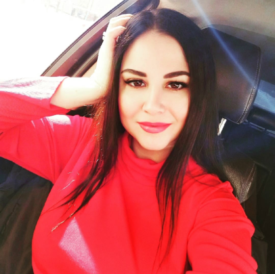 Қыз ұзын қара шашымен қызыл свитерде автомобильде отыр, камераға күлімсіреп жатыр.