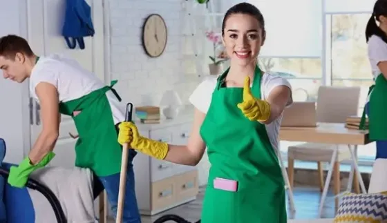Улыбающаяся женщина в зеленом фартуке с желтыми перчатками и веником показывает жест 