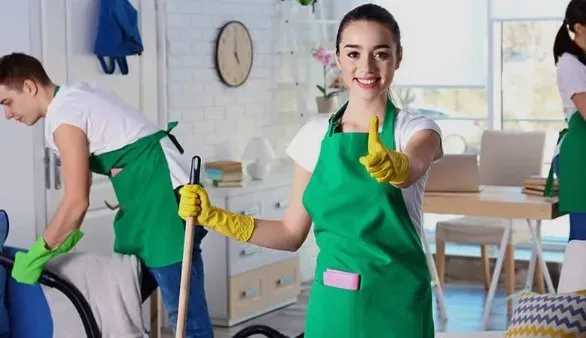 Улыбающаяся женщина в зеленом фартуке с желтыми перчатками и веником показывает жест 