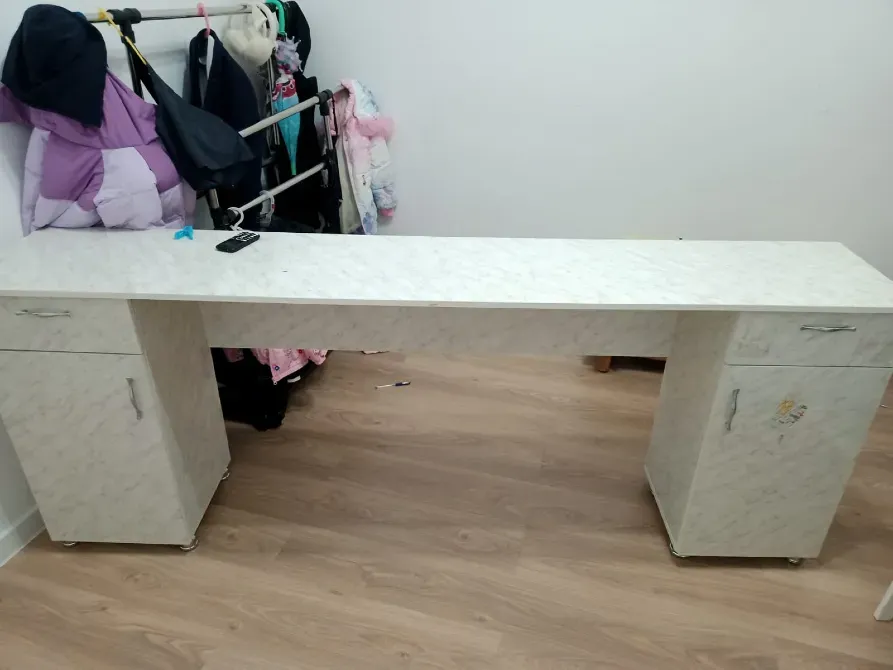 Ақ үстелдің екі бүйірінде орналасқан вертикалды шкафтар, ағаш еденде. Үстелде теледидардың пульті жатыр, ал фонға ілінген заттар көрінеді.