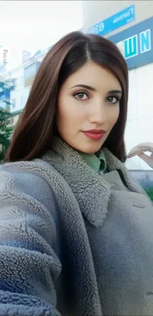 Ұзын шашты және жарқын еріндері бар жас әйел, жайлы шубада киініп, көшеде селфи түсіруде. Фондағы көріністе ғимараттар мен жасыл желектер көрініп тұр.